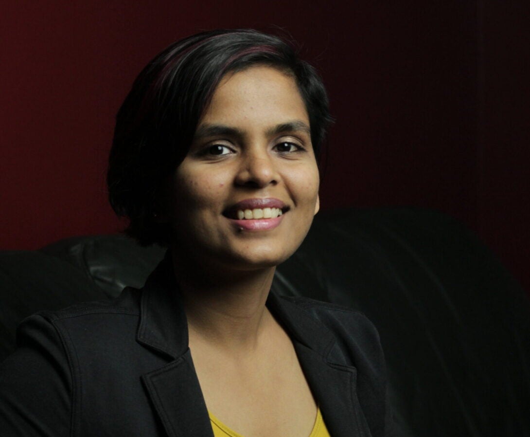 Seated headshot of brown Indian person wearing a black collared blazer over a yellow v-neck top. The person has their fingers interlaced and resting on the armrest of a black leather couch and are smiling at the camera.