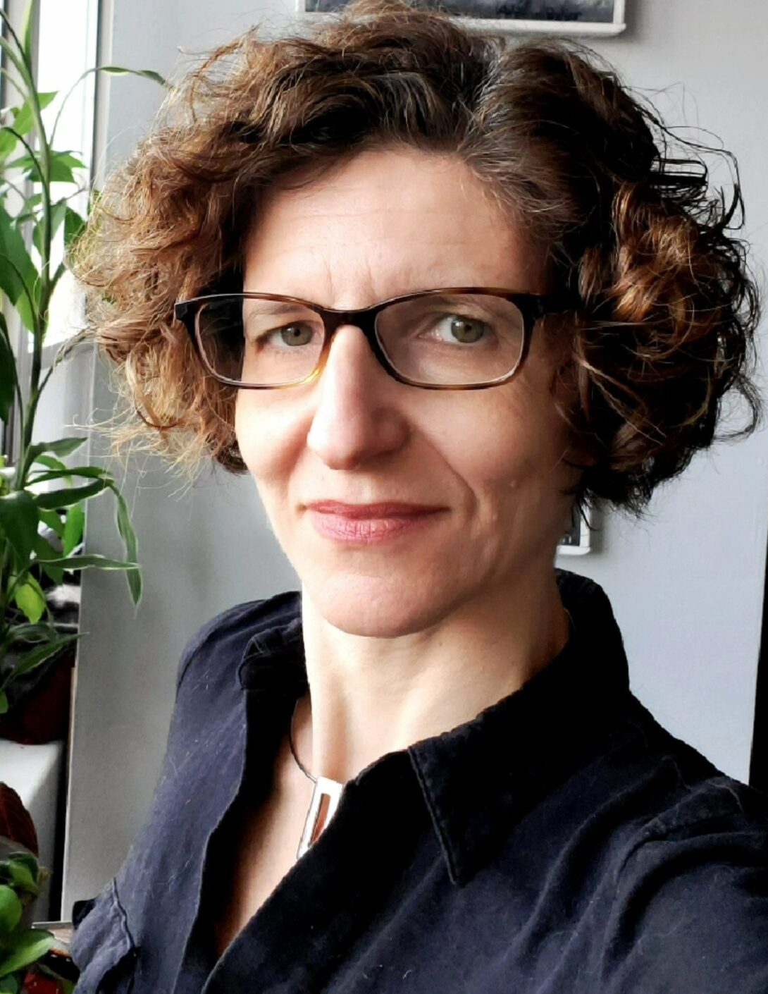 Closeup headshot of white woman with glasses, staring into camera. She has short curly dark brown hair, rectangular rimmed glasses, and is wearing a black collared shirt.