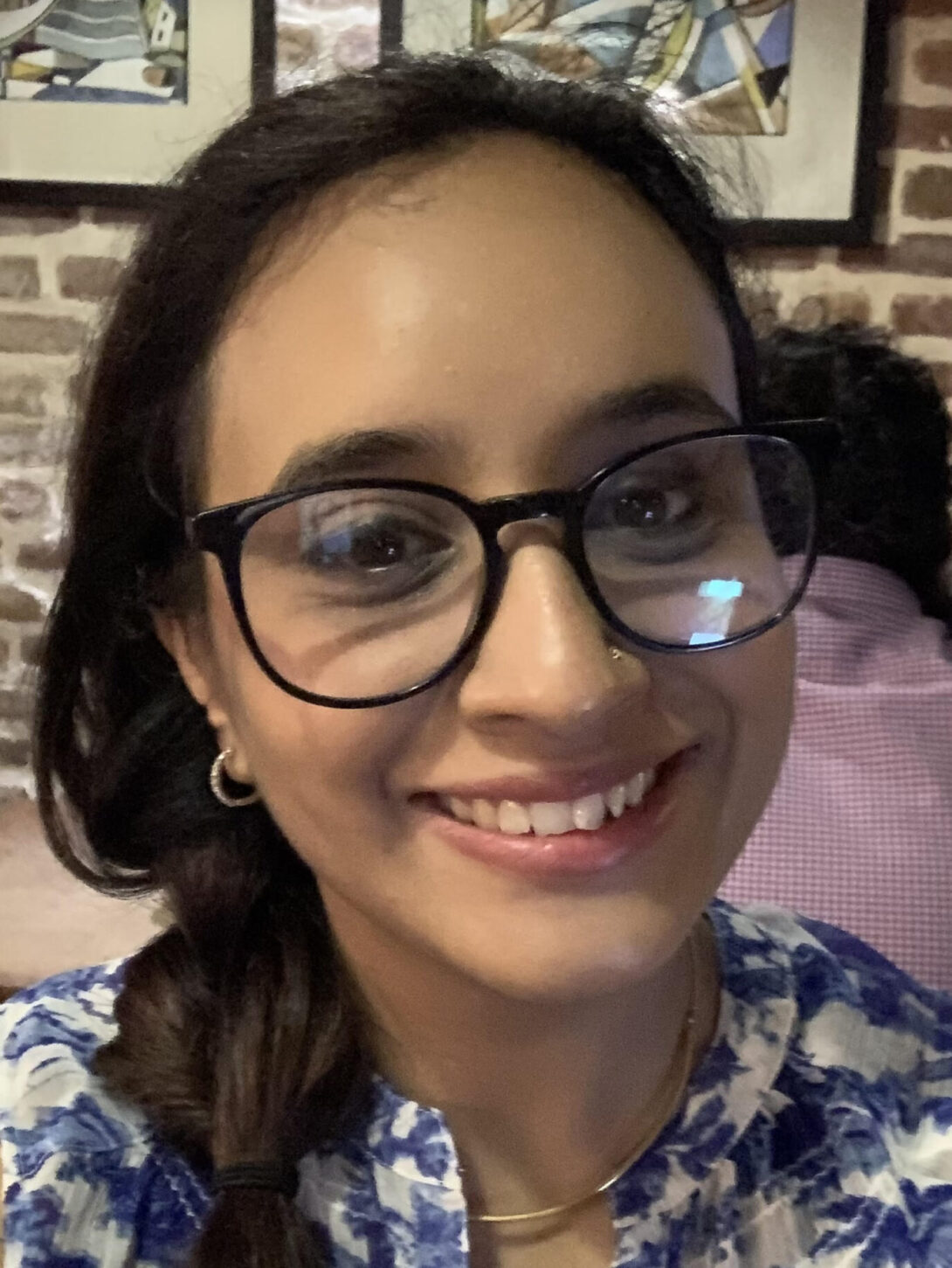 Close up head shot of brown woman with black round-rectangular rimmed glasses, braided hair to the side, wearing a blue and white floral shirt smiling into the camera