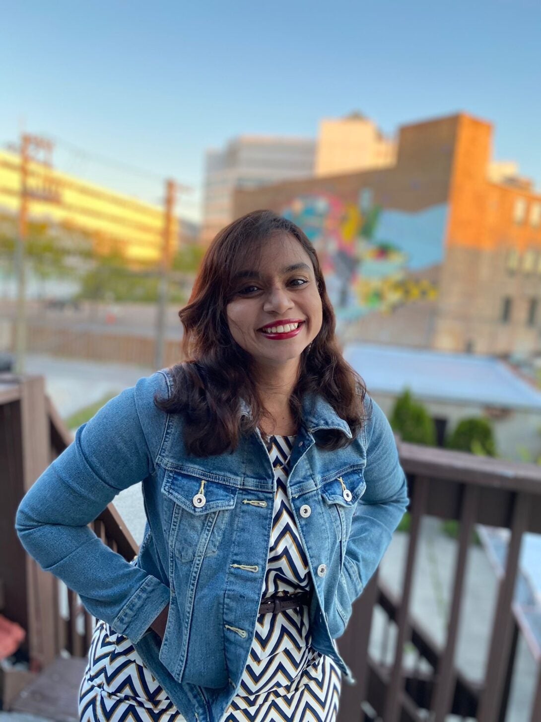 Image of a South Asian woman in a white dress with black stripes and a blue denim jacket, smiling at the camera.